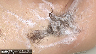 Dark haired woman reclines smiling in muddy tub showing soapy areola on green hill.